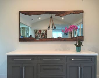 May include: A large, rectangular mirror with a natural wood frame. The frame has a live edge design and is a dark brown color. The mirror is reflecting a room with a chandelier and a painting on the wall. A pink orchid is sitting on a countertop in front of the mirror.