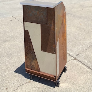 May include: A rusty metal podium with a silver top and wheels. The podium is made of different pieces of metal that have been welded together. The metal has a rustic, industrial look.