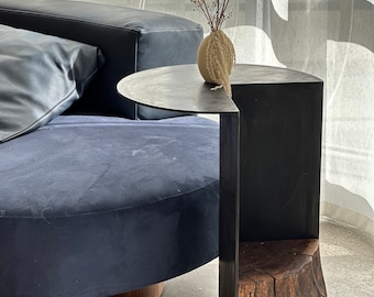 May include: A modern side table with a black metal frame and a wooden base. The table has a half-circle top and a shelf. A small vase with dried flowers sits on the table.