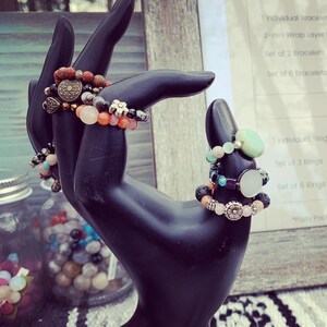 Puede incluir: Un expositor de pulseras y anillos de piedras preciosas de colores en una mano de maniqu&iacute; negra. La lista de precios para pulseras individuales, conjuntos de pulseras, anillos individuales y conjuntos de anillos es visible en el fondo. El texto de la lista de precios dice: "Pulsera individual 15 &euro;", "Pulsera de capa de envoltura 2 en 1 20 &euro;", "Conjunto de 2 pulseras 25 &euro;", "Conjunto de 6 pulseras 60 &euro;", "Anillos", "Anillo individual 6 &euro;", "Conjunto de 3 anillos 15 &euro;", "Conjunto de 6 anillos 25 &euro;", "*Descuentos en paquetes de fiesta disponibles*"