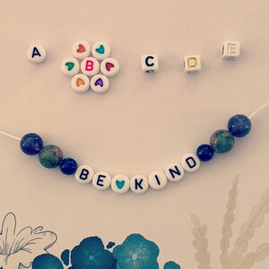 Pu&ograve; includere: Un braccialetto di perline con le parole "BE KIND" scritte con perline di lettere bianche. Il braccialetto presenta perline di pietra blu e verde e una perlina a forma di cuore. Perline di lettere e perline a forma di cuore aggiuntive sono sparse.