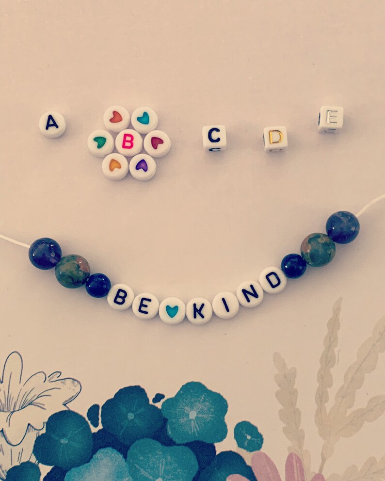 Pu&ograve; includere: Un braccialetto di perline con le parole "BE KIND" scritte con perline di lettere bianche. Il braccialetto presenta perline di pietra blu e verde e una perlina a forma di cuore. Perline di lettere e perline a forma di cuore aggiuntive sono sparse.