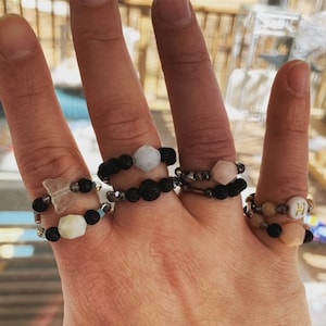 Puede incluir: Una mano lleva cinco anillos hechos de cuentas de roca de lava negra, cuentas plateadas y piedras de varios colores. Las piedras incluyen una mariposa, una piedra azul claro, una piedra rosa, una piedra blanca y una piedra rosa con una letra "H" dorada.