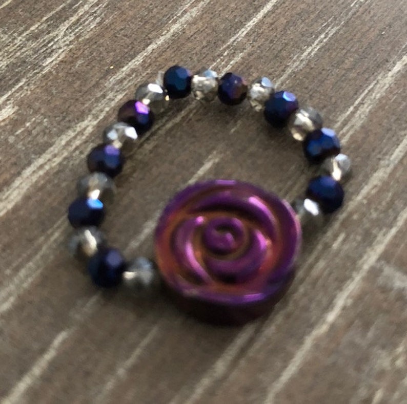 Puede incluir: Un anillo de cuentas de color morado y plateado con un dije de rosa morada.