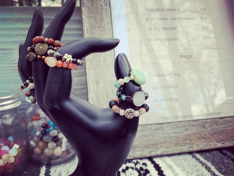 Puede incluir: Un expositor de pulseras y anillos de piedras preciosas de colores en una mano de maniqu&iacute; negra. La lista de precios para pulseras individuales, conjuntos de pulseras, anillos individuales y conjuntos de anillos es visible en el fondo. El texto de la lista de precios dice: "Pulsera individual 15 &euro;", "Pulsera de capa de envoltura 2 en 1 20 &euro;", "Conjunto de 2 pulseras 25 &euro;", "Conjunto de 6 pulseras 60 &euro;", "Anillos", "Anillo individual 6 &euro;", "Conjunto de 3 anillos 15 &euro;", "Conjunto de 6 anillos 25 &euro;", "*Descuentos en paquetes de fiesta disponibles*"