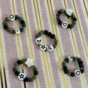 May include: Five beaded rings with various white charms, including a heart, moon, star, and flower. The rings feature a mix of dark green, purple, and black beads. The background is a pink and gold striped pattern.
