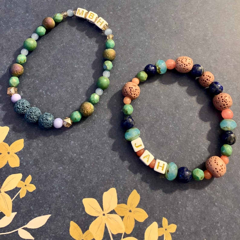 Pu&ograve; includere: Due braccialetti di perline colorate. Uno mostra le lettere "MBH" su perline bianche, l'altro "JAH". Le perline sono verdi, blu, marroni e rosa, con diverse texture. Un accessorio di moda vivace e originale.