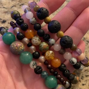 Pu&ograve; includere: Diversi braccialetti di perline con una variet&agrave; di pietre preziose colorate e perline di lava nere. I braccialetti presentano perline verdi, arancioni, rosa e viola, infilate su cordoncini elastici. I braccialetti sono tenuti in una mano.