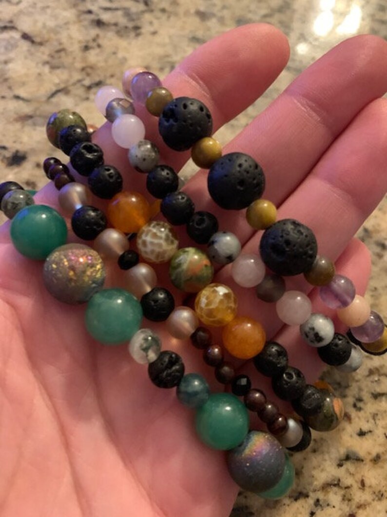 Pu&ograve; includere: Diversi braccialetti di perline con una variet&agrave; di pietre preziose colorate e perline di lava nere. I braccialetti presentano perline verdi, arancioni, rosa e viola, infilate su cordoncini elastici. I braccialetti sono tenuti in una mano.
