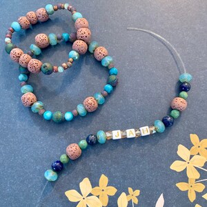 Pu&ograve; includere: Tre braccialetti di perline e una collana con la parola "JAH". I braccialetti presentano perline marroni, turchesi e verdi. La collana ha perline simili, con le lettere "JAH" scritte in perline quadrate bianche.