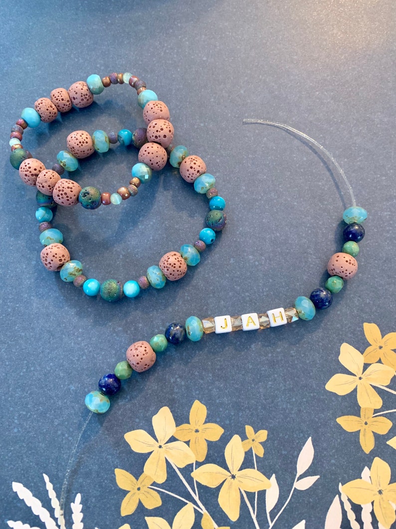 Pu&ograve; includere: Tre braccialetti di perline e una collana con la parola "JAH". I braccialetti presentano perline marroni, turchesi e verdi. La collana ha perline simili, con le lettere "JAH" scritte in perline quadrate bianche.