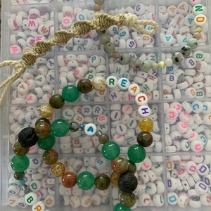 Pu&ograve; includere: Una collezione di braccialetti di perline con vari colori e trame. Un braccialetto dice "HEY FRIEND" in perline di lettere colorate. Un altro braccialetto dice "BREATHE". I braccialetti presentano perline verdi, marroni e nere.