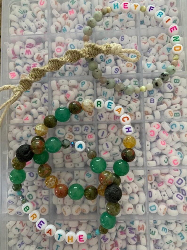 Pu&ograve; includere: Una collezione di braccialetti di perline con vari colori e trame. Un braccialetto dice "HEY FRIEND" in perline di lettere colorate. Un altro braccialetto dice "BREATHE". I braccialetti presentano perline verdi, marroni e nere.