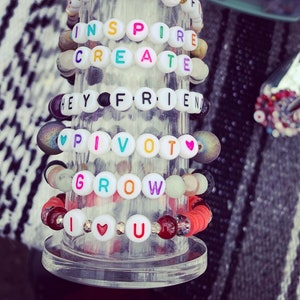 May include: A collection of beaded bracelets with white letter beads spelling out inspirational words like "INSPIRE", "CREATE", "HEY FRIEND", "PIVOT", "GROW", and "I LOVE U". The bracelets feature various colors and bead styles.