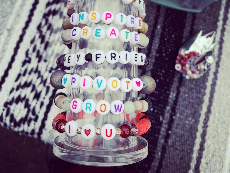Pu&ograve; includere: Una collezione di braccialetti di perline con perline di lettere bianche che compongono parole ispiratrici come "INSPIRE", "CREATE", "HEY FRIEND", "PIVOT", "GROW" e "I LOVE U". I braccialetti presentano vari colori e stili di perline.