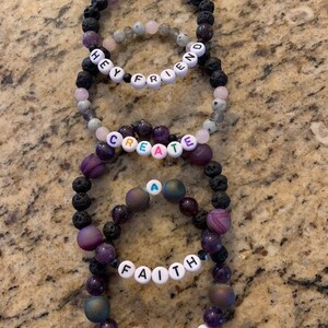 Pu&ograve; includere: Diversi braccialetti di perline con perline viola, nere e rosa sono impilati su una superficie strutturata. Alcuni braccialetti hanno perline di lettere bianche che compongono le parole "HEY FRIEND", "CREATE", "FAITH" e "PIVOT".