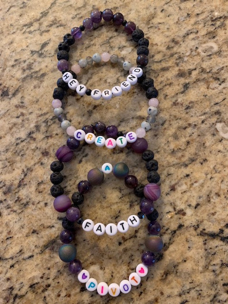 Pu&ograve; includere: Diversi braccialetti di perline con perline viola, nere e rosa sono impilati su una superficie strutturata. Alcuni braccialetti hanno perline di lettere bianche che compongono le parole "HEY FRIEND", "CREATE", "FAITH" e "PIVOT".