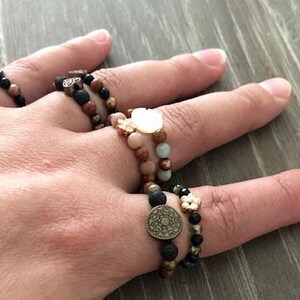 Puede incluir: Una mano con varios anillos de cuentas con detalles met&aacute;licos. Los anillos est&aacute;n hechos de roca volc&aacute;nica negra, madera marr&oacute;n y piedras marr&oacute;n claro. Los anillos est&aacute;n apilados uno encima del otro y se llevan en el dedo medio.