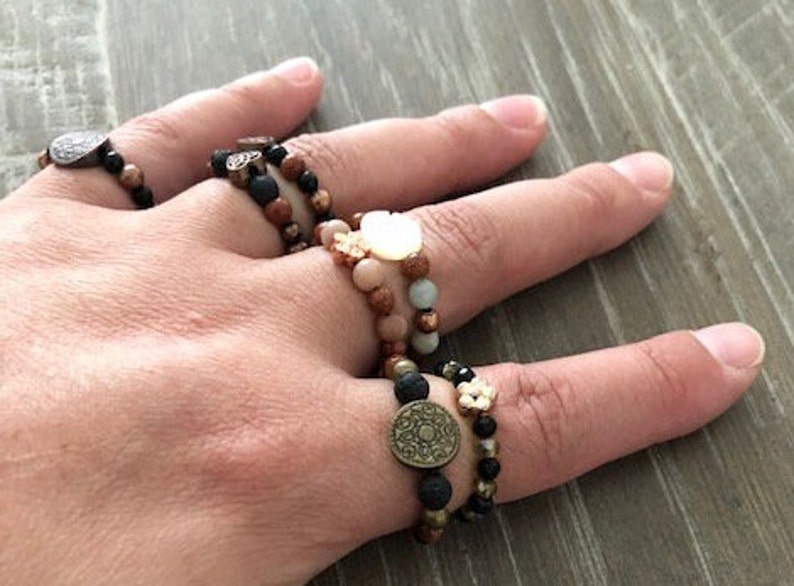 Puede incluir: Una mano con varios anillos de cuentas con detalles met&aacute;licos. Los anillos est&aacute;n hechos de roca volc&aacute;nica negra, madera marr&oacute;n y piedras marr&oacute;n claro. Los anillos est&aacute;n apilados uno encima del otro y se llevan en el dedo medio.