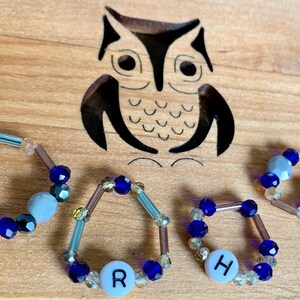 Puede incluir: Cuatro anillos de cuentas con cuentas azules y transparentes, cada uno con una cuenta de letra blanca. Las letras deletrean la palabra "R-O-H-H".