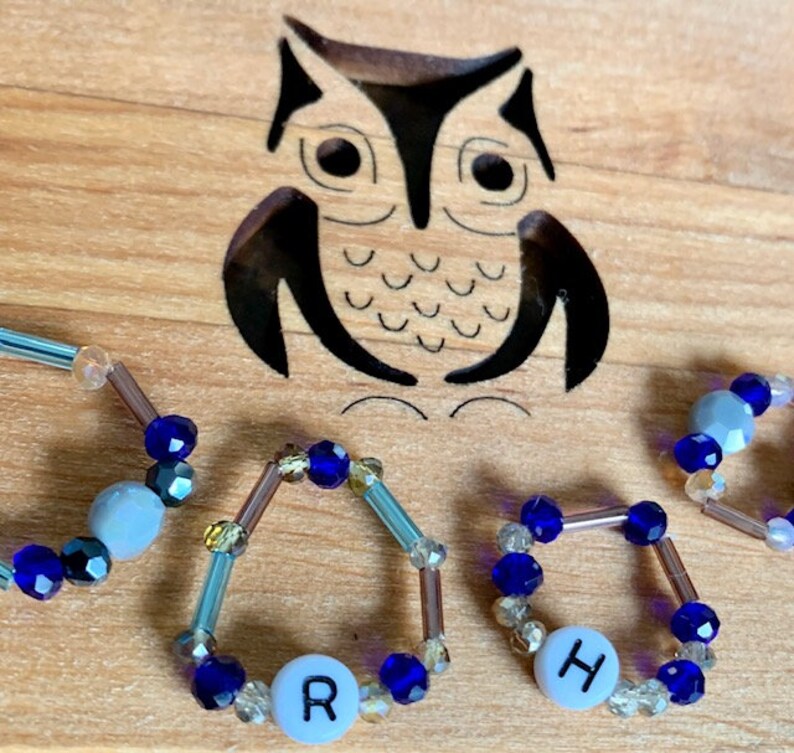 Puede incluir: Cuatro anillos de cuentas con cuentas azules y transparentes, cada uno con una cuenta de letra blanca. Las letras deletrean la palabra "R-O-H-H".