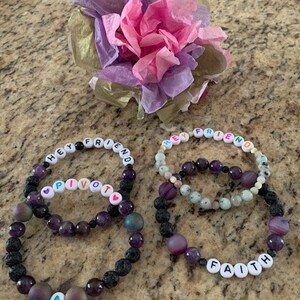 Pu&ograve; includere: Due braccialetti di perline con le parole "HEY FRIEND" e "PIVOT" scritte con perline di lettere bianche. I braccialetti presentano perline viola e nere. Un terzo braccialetto dice "FAITH". Un fiore di carta decorativo &egrave; sullo sfondo.