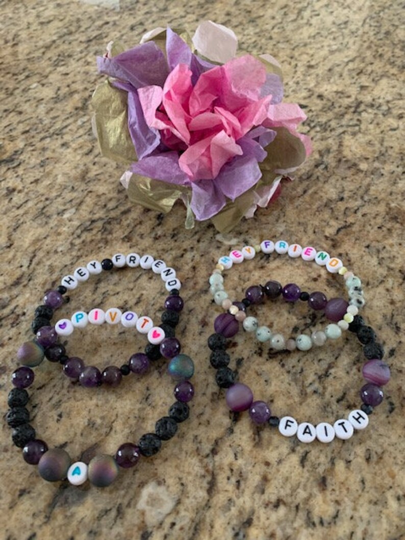 Pu&ograve; includere: Due braccialetti di perline con le parole "HEY FRIEND" e "PIVOT" scritte con perline di lettere bianche. I braccialetti presentano perline viola e nere. Un terzo braccialetto dice "FAITH". Un fiore di carta decorativo &egrave; sullo sfondo.