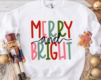 May include: White sweatshirt with the text "Merry and Bright" in colorful, handwritten letters. The sweatshirt is on a wooden surface with a nutcracker and Christmas ornaments.
