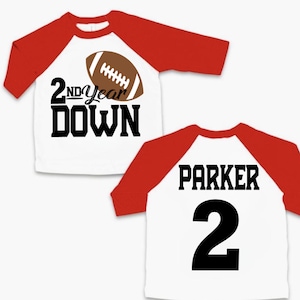 May include: Two white and red baseball shirts with a football design. The first shirt says "2nd Year Down" with a football graphic. The second shirt says "Parker 2".