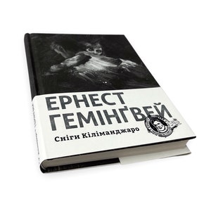 以下が含まれることがあります： ハードカバーの本で、表紙に白黒のイラストが描かれています。タイトルと著者の名前は、白い帯に太字の黒い文字で印刷されています。テキストは「アーネスト・ヘミングウェイ」と「キリマンジャロの雪」です。