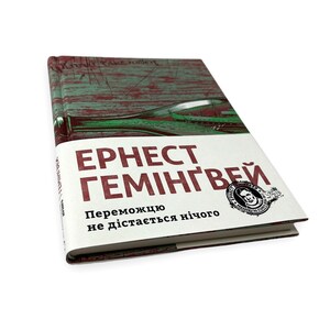 以下が含まれることがあります： 緑と赤の背景にボトルと「ERNEST HEMINGWAY」と「Переможцю не дістається нічого」の文字が書かれた本の表紙です。