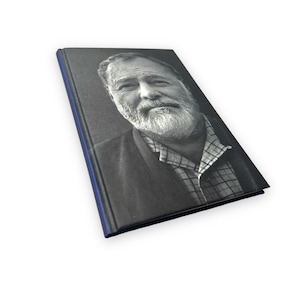 以下が含まれることがあります： 本の表紙に、ひげと口ひげを生やした男性の白黒写真が写っています。本は濃い青色の背表紙です。