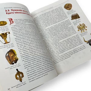 May include: Open book with a gold-colored metal cup and a gold-colored metal brooch on the left page. The right page features text about the history of metalwork in the Minoan civilization. The text includes the words "Minoan", "metalwork", and "history".