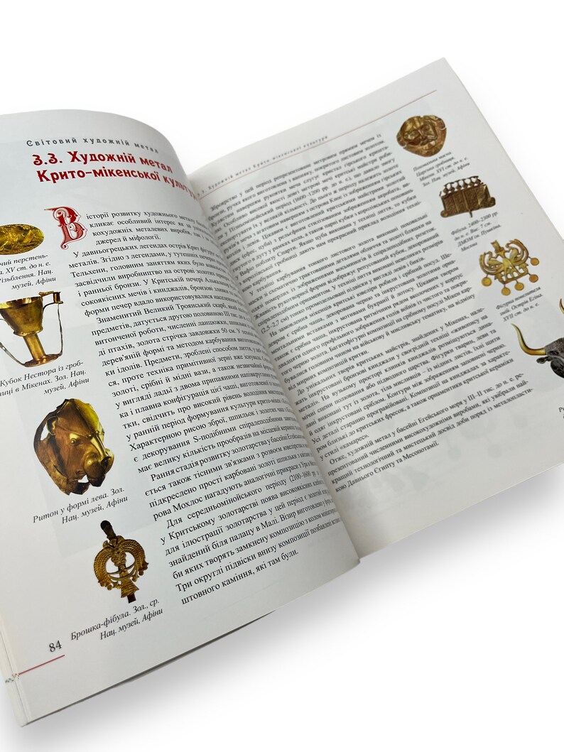 May include: Open book with a gold-colored metal cup and a gold-colored metal brooch on the left page. The right page features text about the history of metalwork in the Minoan civilization. The text includes the words "Minoan", "metalwork", and "history".