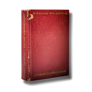 May include: A vintage book with a red cover and gold lettering. The title "РОМАН ФЕДОРІВ" is at the top, and "ОПТІЧНИЙ СВІТИЛЬНИК" is on the spine and bottom. The cover has a subtle embossed design.