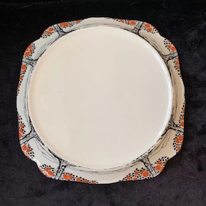 May include: A white ceramic plate with a scalloped edge and a black and orange design. The design features stylised trees with orange circles.