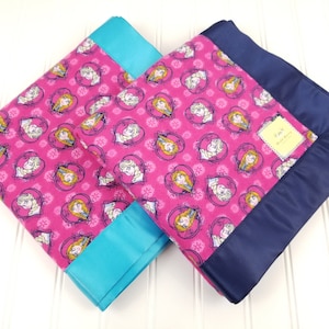 May include: Two pink fleece blankets with a Frozen character print. One blanket has a turquoise satin trim and the other has a navy blue satin trim.