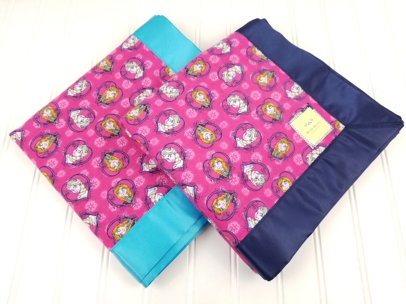 May include: Two pink fleece blankets with a Frozen character print. One blanket has a turquoise satin trim and the other has a navy blue satin trim.