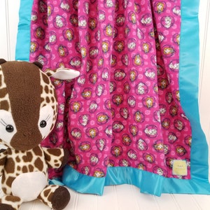 May include: A pink fleece baby blanket with a repeating pattern of cartoon girls' faces in hearts. The blanket has a turquoise satin border.