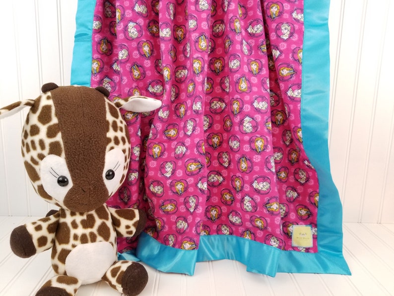 May include: A pink fleece baby blanket with a repeating pattern of cartoon girls' faces in hearts. The blanket has a turquoise satin border.