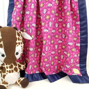 May include: A pink fleece baby blanket with a navy blue satin border. The blanket features a repeating pattern of hearts with images of princesses inside. A brown and tan stuffed giraffe is sitting in front of the blanket.