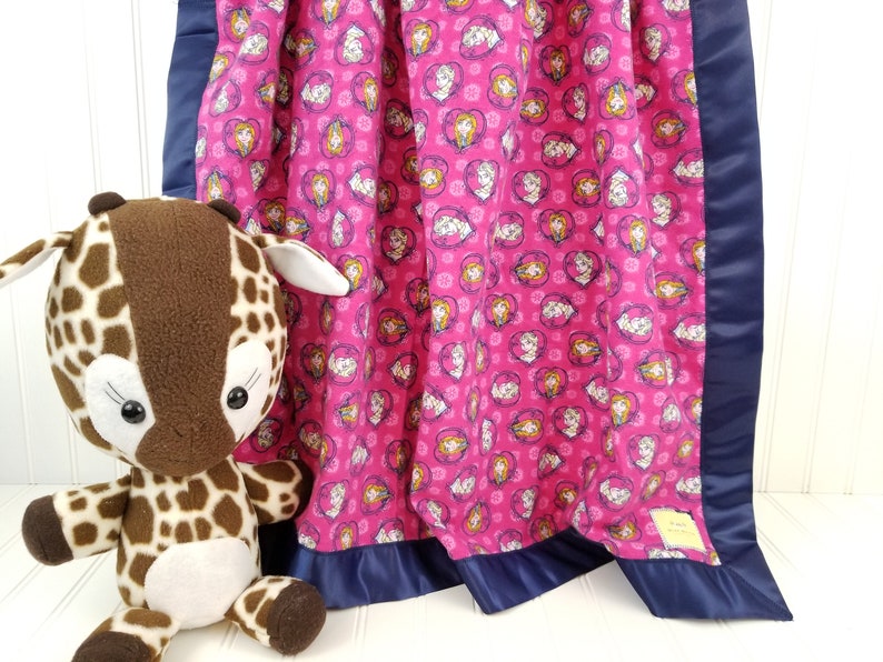 May include: A pink fleece baby blanket with a navy blue satin border. The blanket features a repeating pattern of hearts with images of princesses inside. A brown and tan stuffed giraffe is sitting in front of the blanket.
