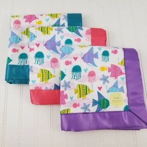 May include: Three baby blankets with a colorful sea life print. The blankets have a satin trim in teal, pink, and purple.