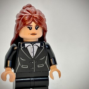 May include: A Lego minifigure of a woman with brown hair wearing a black suit jacket and pants. She has a white shirt underneath the jacket.