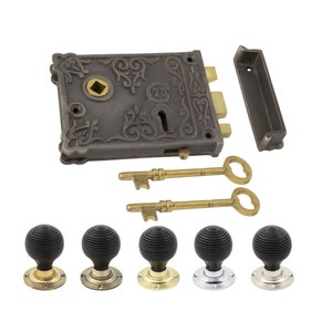 May include: An antique-style door lock set. It features a dark grey metal lock plate with an ornate design, two brass keys, a strike plate, and five black door knobs with brass, chrome, and nickel bases. The lock plate has an ornate design.