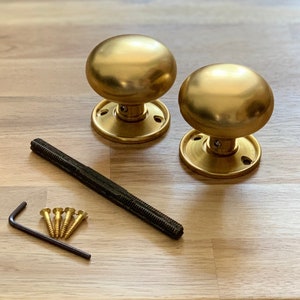 May include: Two brass door knobs with a round design and matching base plates. The hardware set includes a threaded rod, screws, and an Allen key. The knobs have a polished finish and are displayed on a light wood surface.
