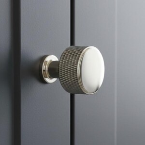 May include: A silver-toned cabinet knob with a textured cylindrical body and a smooth, circular face. The knob is mounted on a dark grey cabinet door. The design is modern and minimalist, suitable for various interior styles.