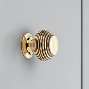 May include: A brass cabinet knob with a ribbed, spherical design. The knob is mounted on a light grey surface. The knob has a circular base and a smooth, flat end. Suitable for cabinets and drawers.