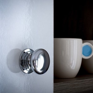 May include: A polished chrome door knob on a white door. Two white ceramic mugs with coloured handles, one blue and one orange, sit on a dark shelf. The mugs are on the right.