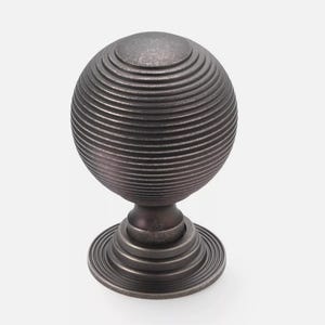 May include: A bronze-coloured cabinet knob with a spherical design. The knob has a series of concentric, horizontal ridges, creating a textured surface. It sits on a circular base with similar ridged detailing, suitable for furniture.